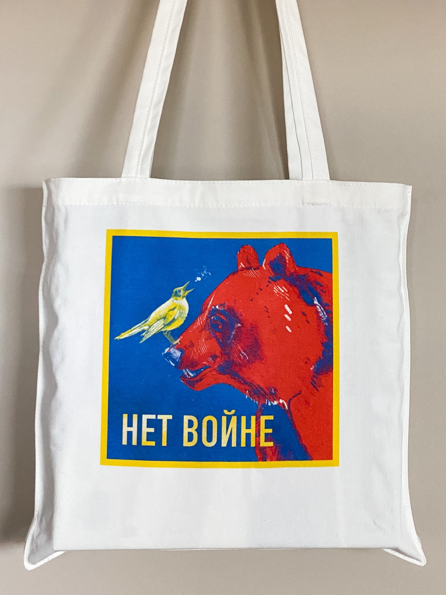 Cotton canvas tote bag depicting an illustration of a yellow nightingale bird looking up and to the right, singing, while standing on the nose of a big red bear. Text says Нет войне.