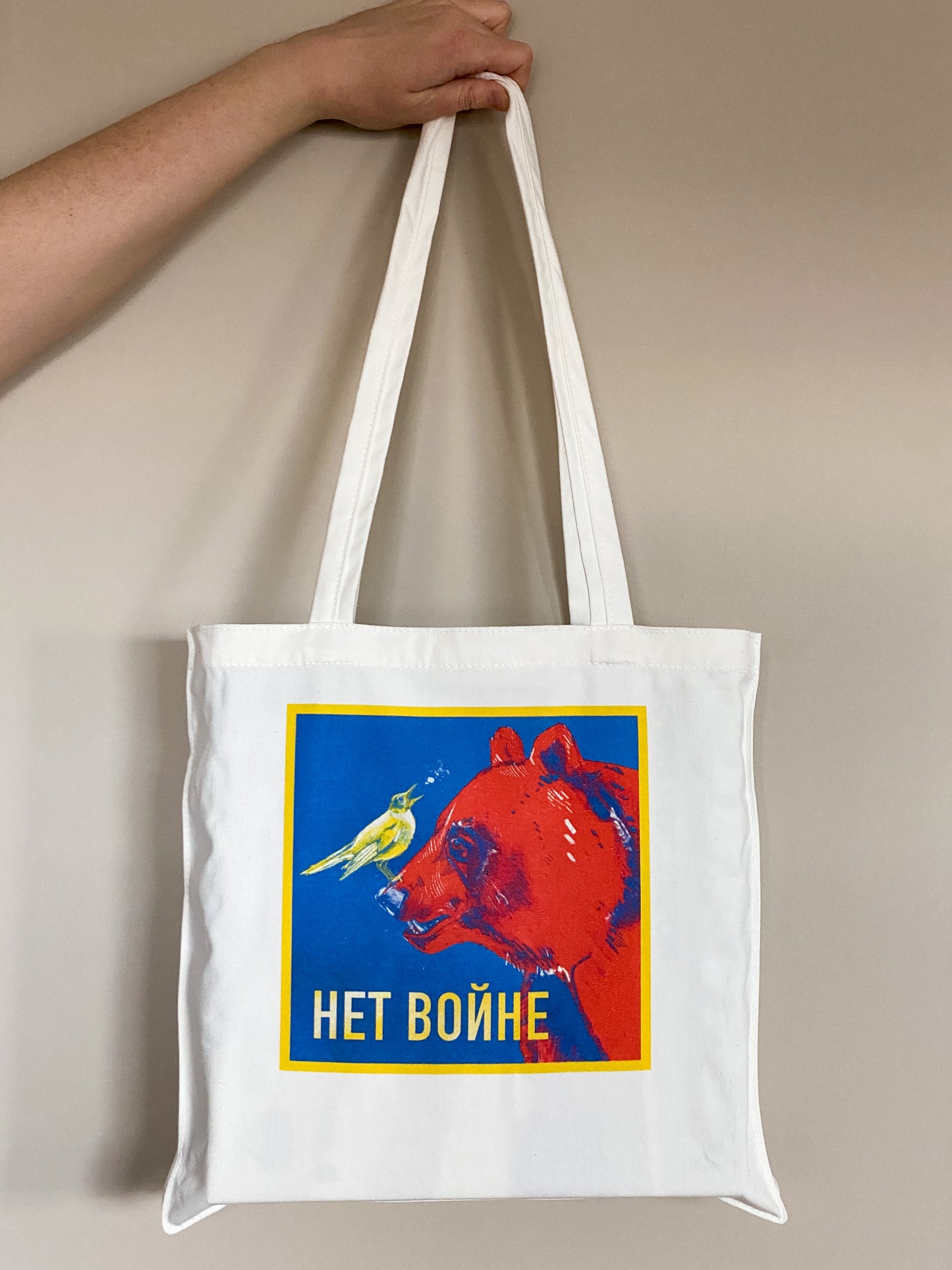 Cotton canvas tote bag depicting an illustration of a yellow nightingale bird looking up and to the right, singing, while standing on the nose of a big red bear. Text says Нет войне.