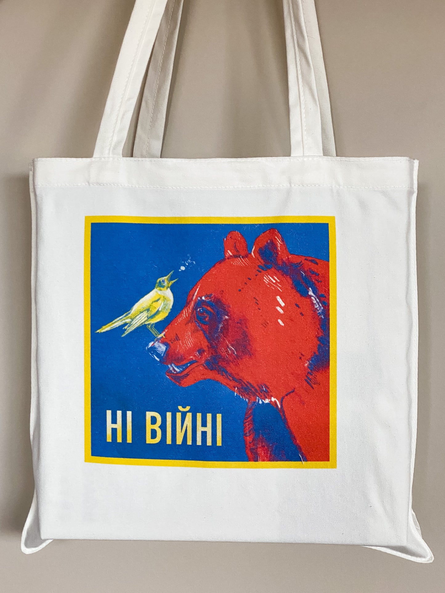 Cotton canvas tote bag depicting an illustration of a yellow nightingale bird looking up and to the right, singing, while standing on the nose of a big red bear. Text says ні війні.