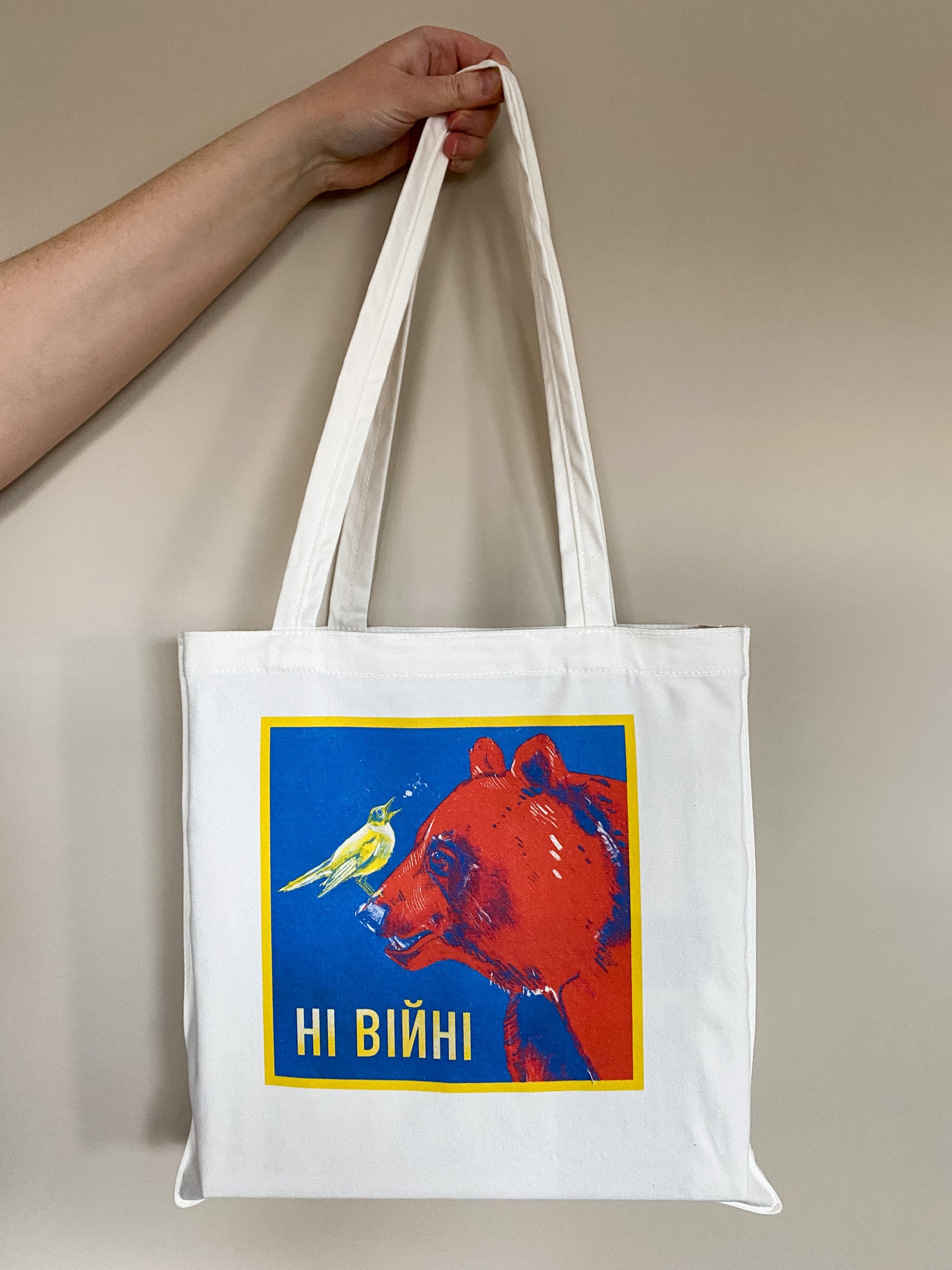 Cotton canvas tote bag depicting an illustration of a yellow nightingale bird looking up and to the right, singing, while standing on the nose of a big red bear. Text says ні війні.