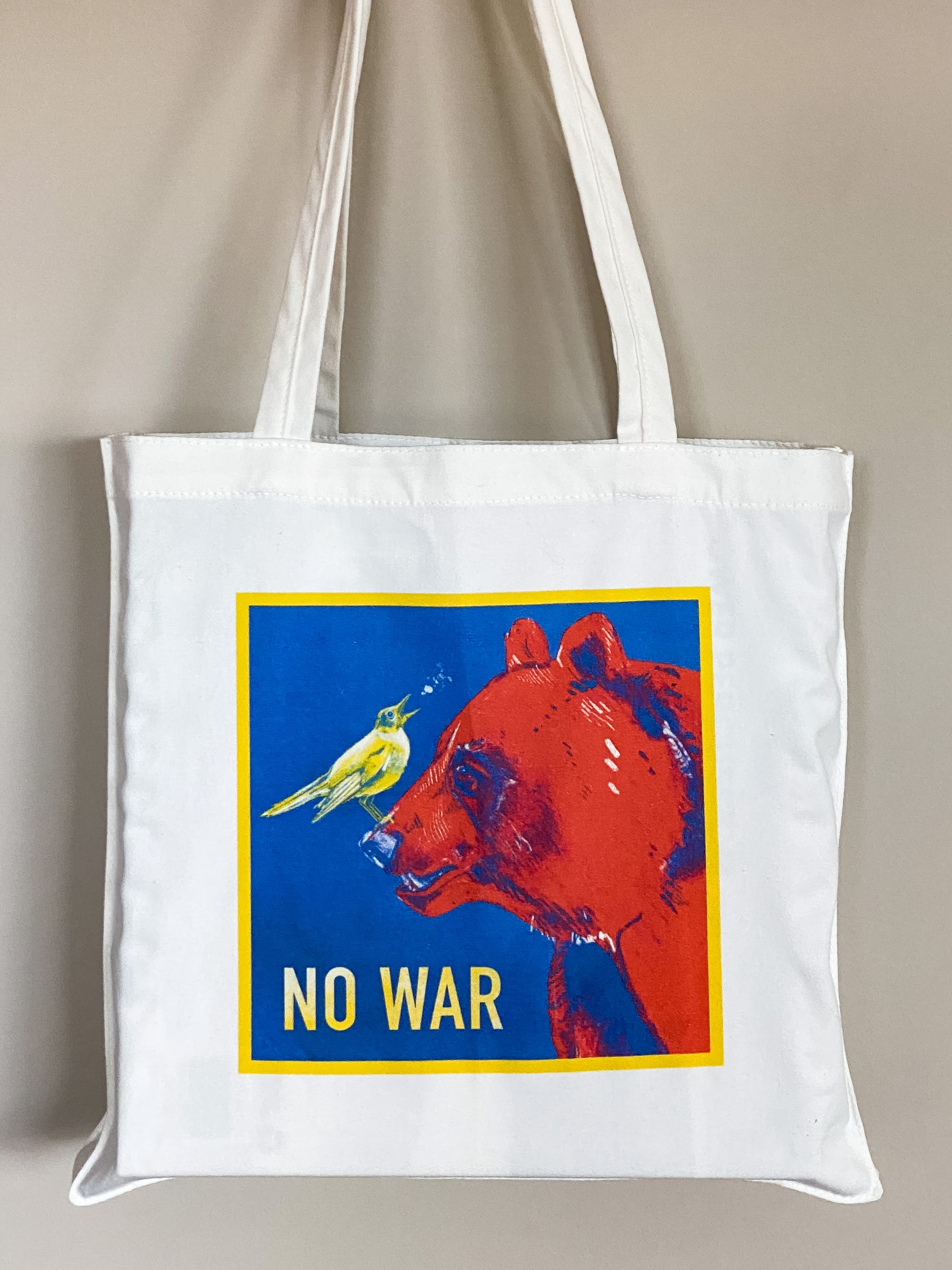 Cotton canvas tote bag depicting an illustration of a yellow nightingale bird looking up and to the right, singing, while standing on the nose of a big red bear. Text says No War.