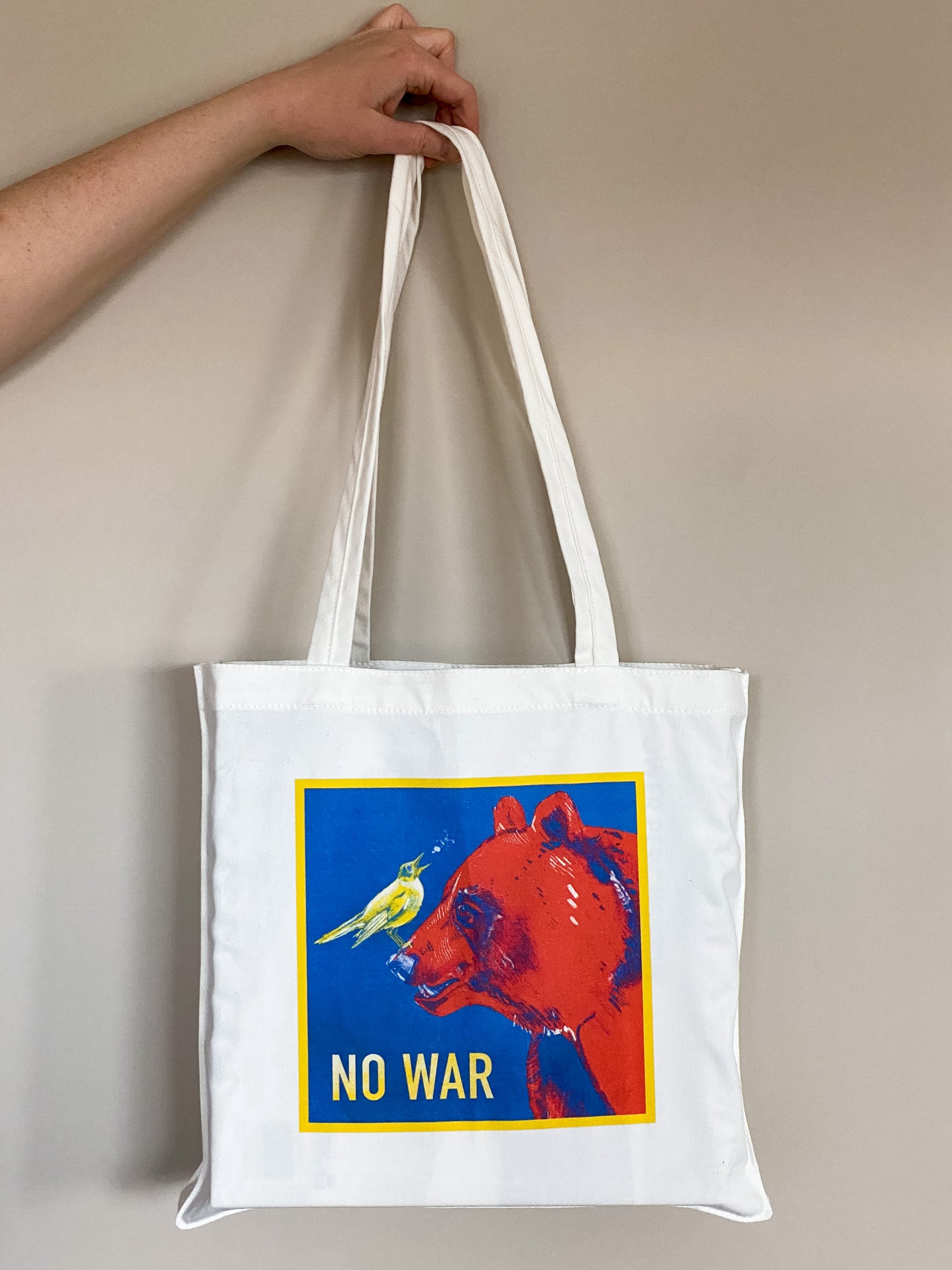 Cotton canvas tote bag depicting an illustration of a yellow nightingale bird looking up and to the right, singing, while standing on the nose of a big red bear. Text says No War.