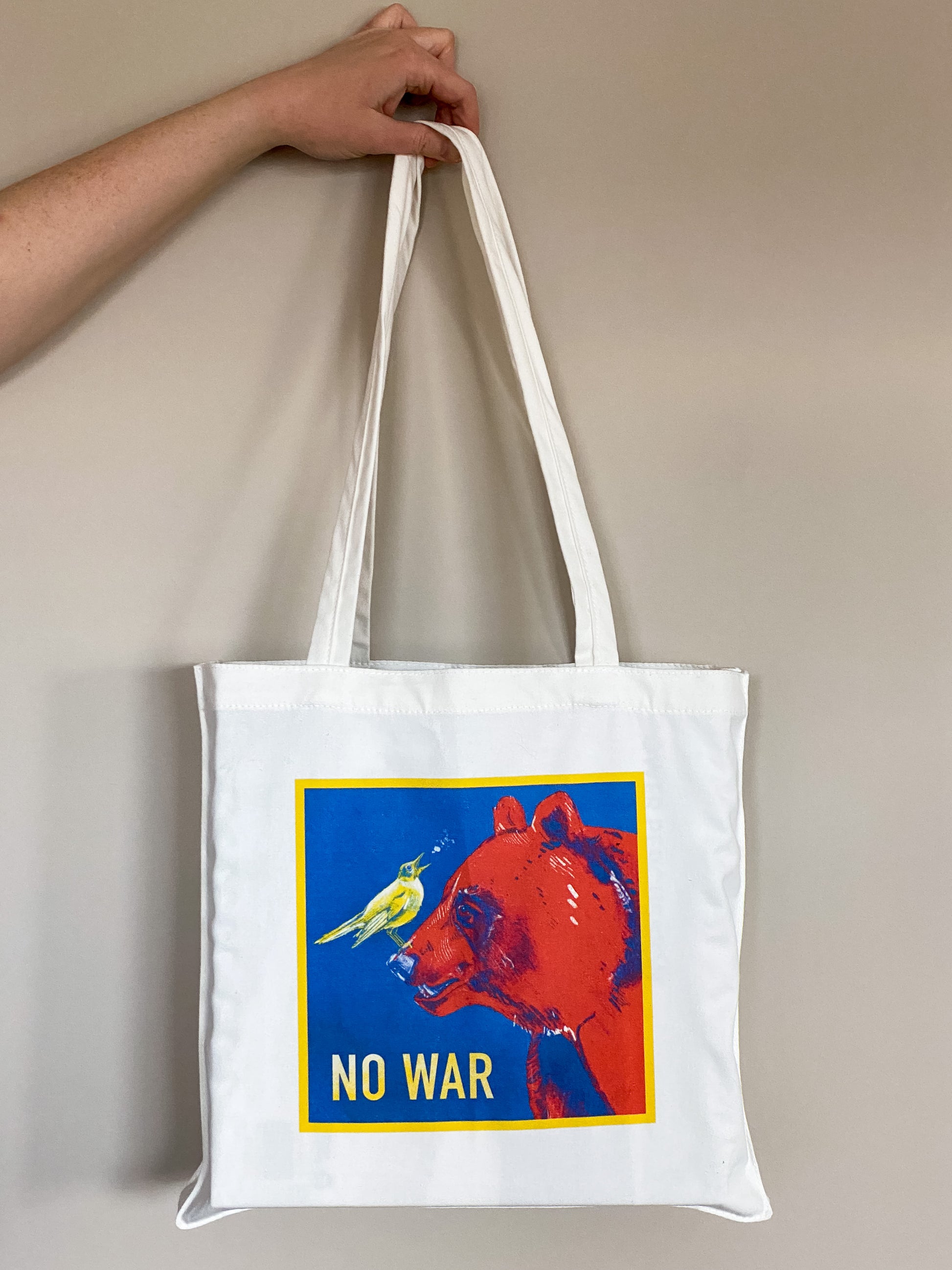 Cotton canvas tote bag depicting an illustration of a yellow nightingale bird looking up and to the right, singing, while standing on the nose of a big red bear. Text says No War.